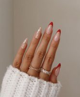 Mão branca com manga branca, anéis e unha vermelha com francesinha e brilho dourado