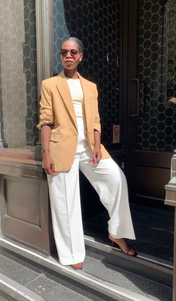 Mulher negra de aproximadamente 40 anos. Ela está usando blazer e calça de alfaiataria com salto alto.