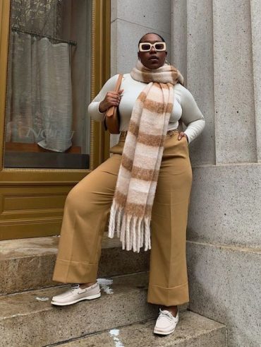 Mulher negra e alta. Ela usa calça caqui, blusa off white e cachecol xadrez