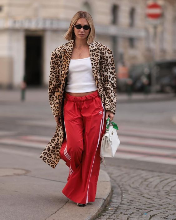 Mulher magra e alta de cabelos curtos e claros usando casaco animal print de onça e calça adidas vermelha