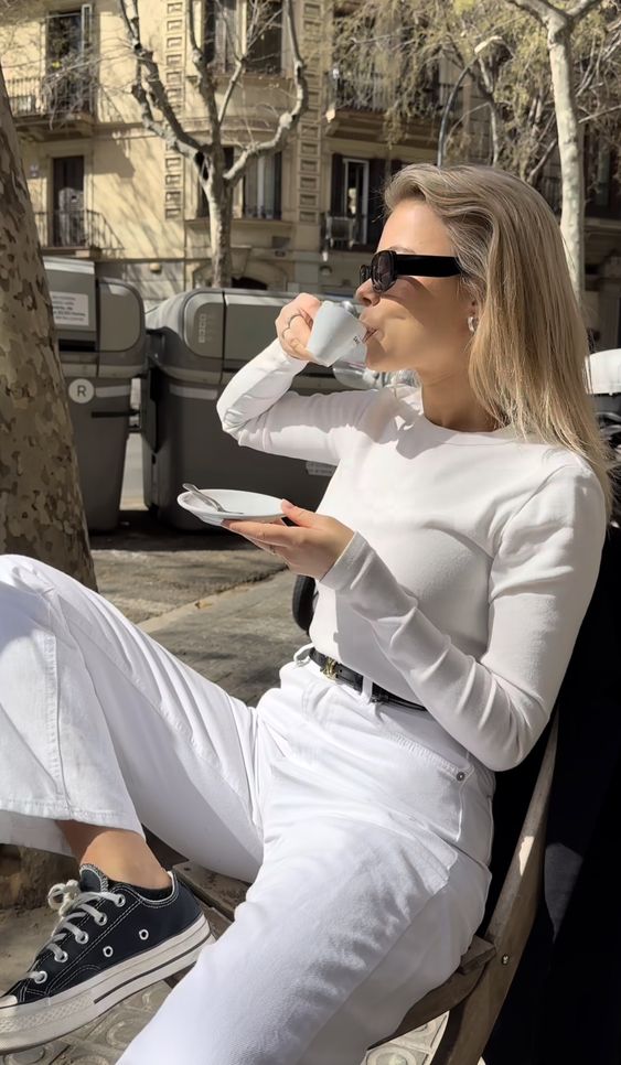 Mulher branca sentada em uma cadeira com uma perna para cima. Ela usa all star preto e blusa branca e está segurando uma xícara