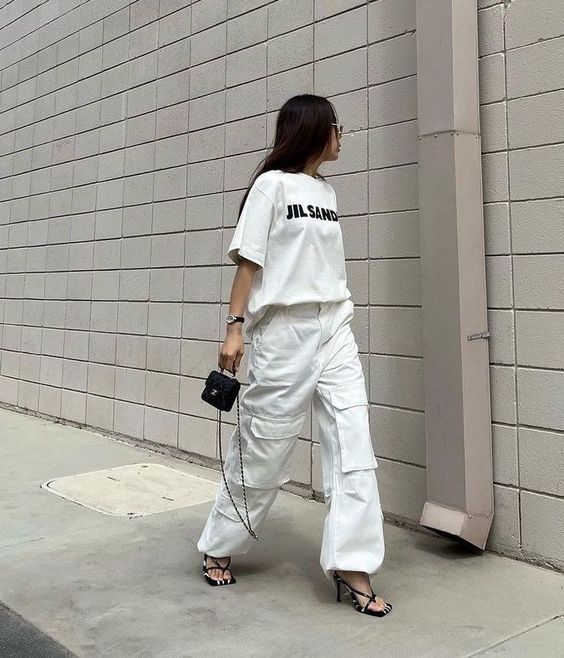 Mulher usando camiseta branca e calça branca larga. Ela segura uma bolsa pequena preta.
