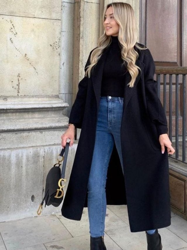 Mulher loira de calça jeans, sobretudo preto, bota de cano baixo e preta, segurando uma bolsa