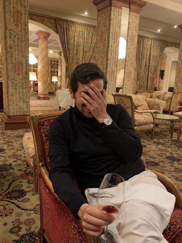 Homem branco com a mão no rosto. Ele está sentado, usando blusa preta e calça branca, com uma taça de vinho na mão