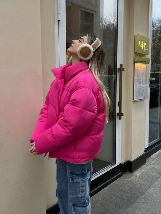 Mulher loira de pele clara usando jaqueta puffer rosa. Ela está olhando para cima