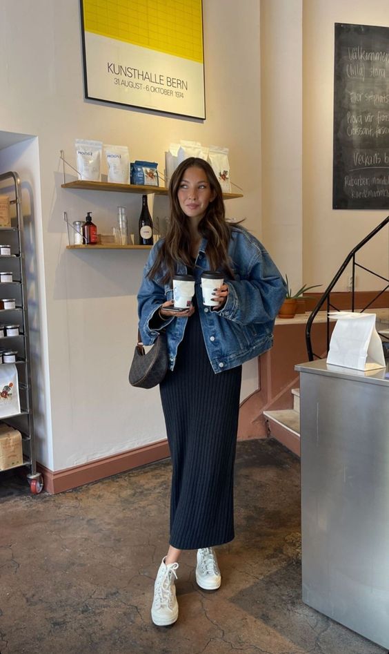 Mulher branca e cabelos claros e lisos. Ela usa jaqueta jeans e vestido longo preto com tenis.