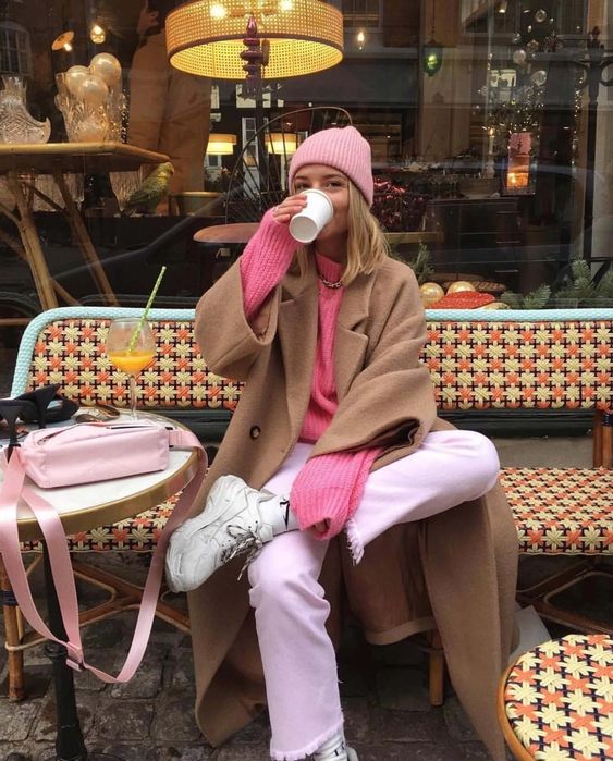 Mulher tomando café em frente a uma rua, ela usa look rosa e tênis branco0