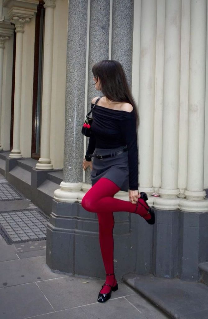Moça branca e magra. Ela usa blusa preta, saia de alfaiatatria, a tendência meia-calça vermelha e calçado preto