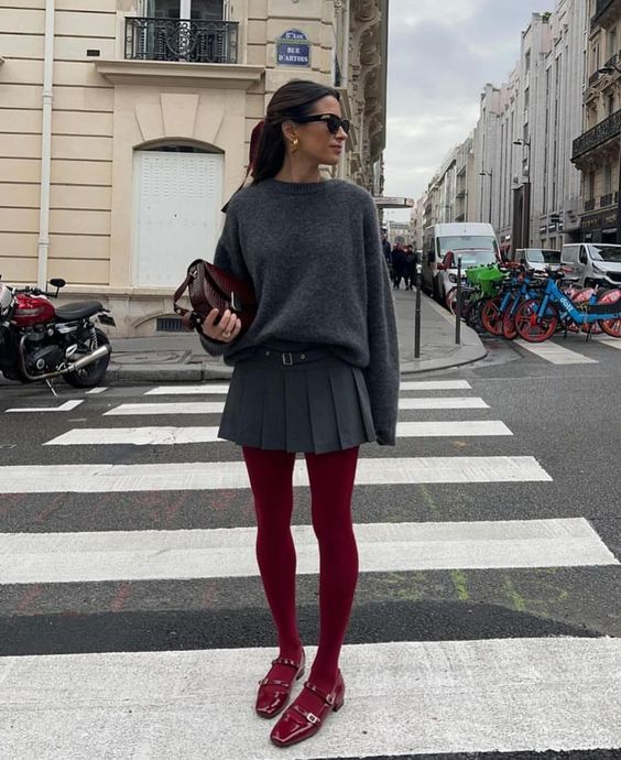 Mulher jovem e magra está de oculos escuros segurando uma bolsa no meio da rua; ela veste suéter cinza e saia cinza plissada com meia-calça vermelha e sapato boneca.