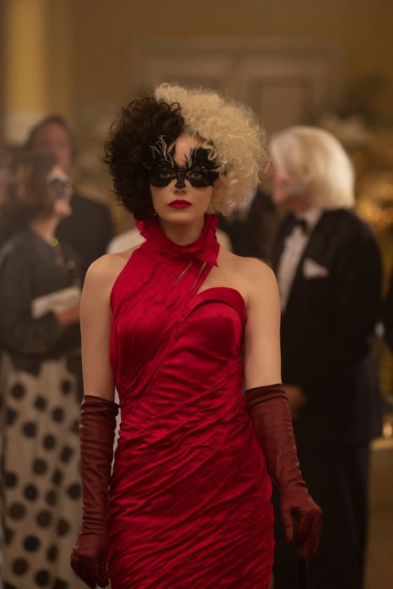 Emma Stone como Cruella. Ela usa máscara preta no rosto, vestido vermelho e luvas bordô