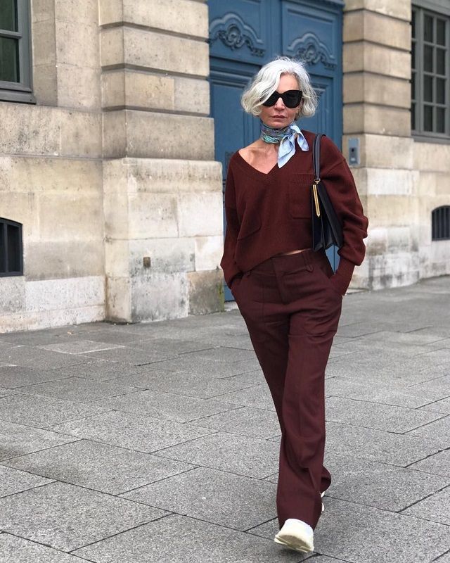 Grece Ghanem usando lenço no pescoço, tricô marrom, calça de alfaiataria marrom e sapato branco