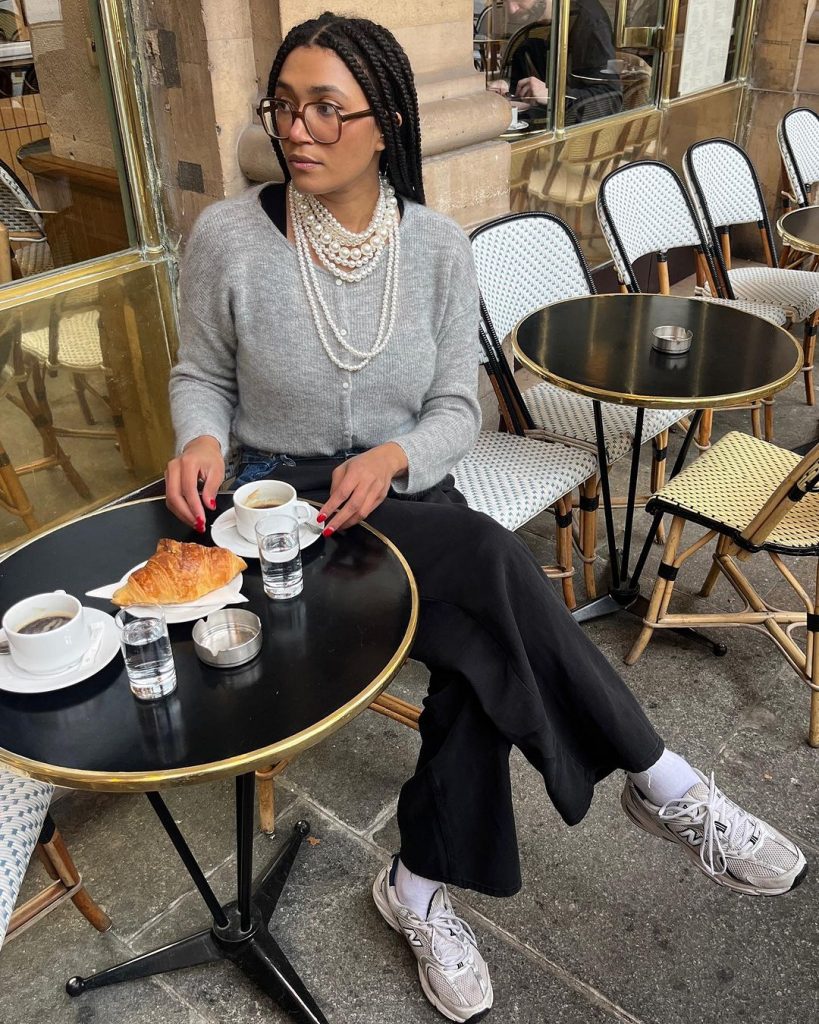 Lena Farouil usando colar de pérolas, suéter cinza, calça jeans e tênis New Balance