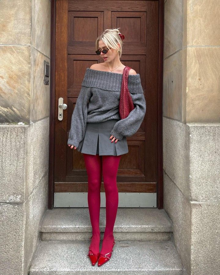 Linda usando tricô cinza ombro a ombro, minissaia plissada cinza, meia-calça vermelho cereja e slingback Miu Miu