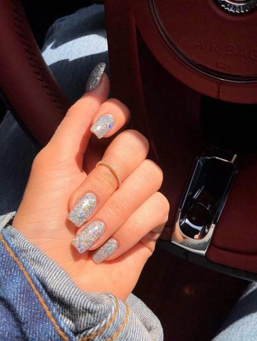 Mão branca curvada com anel e unha da Kylie Jenner com brilhosas grossos