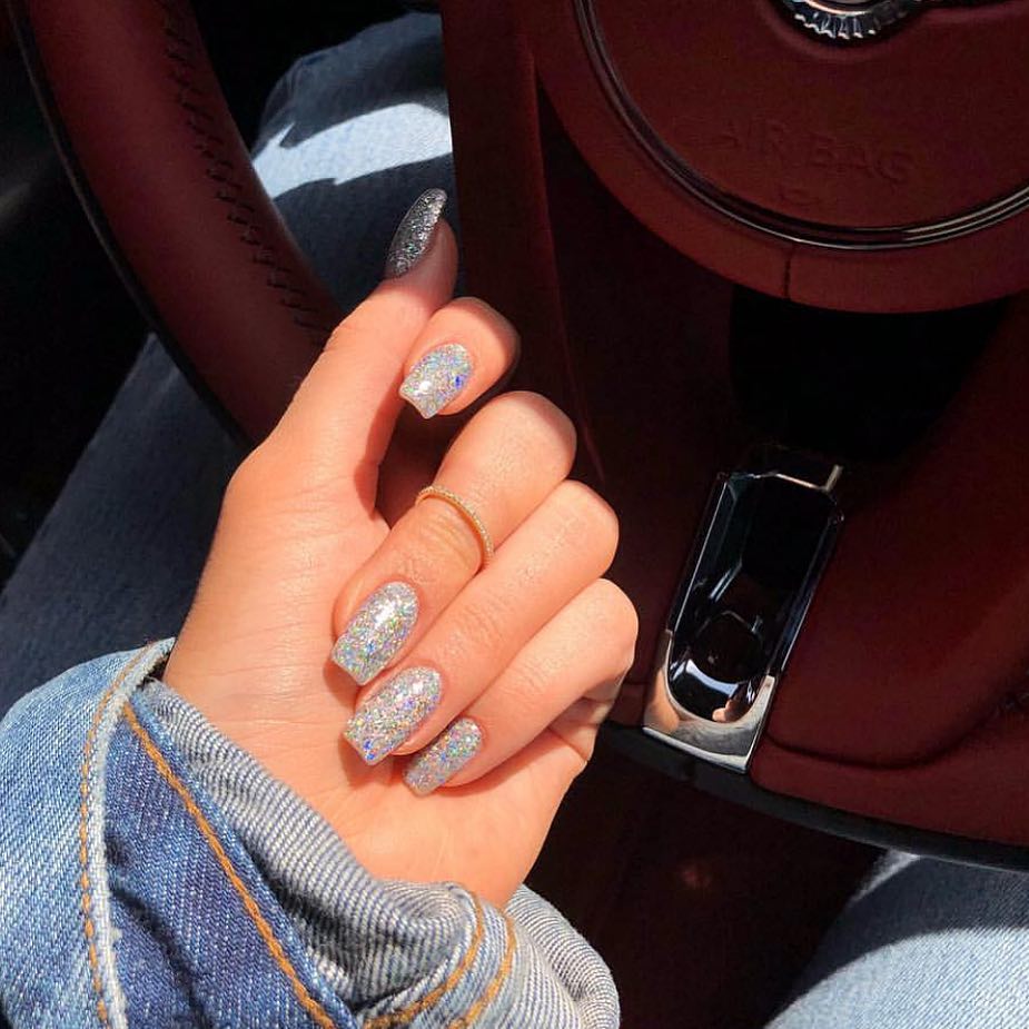Mão branca curvada com anel e unha da Kylie Jenner com brilhosas grossos