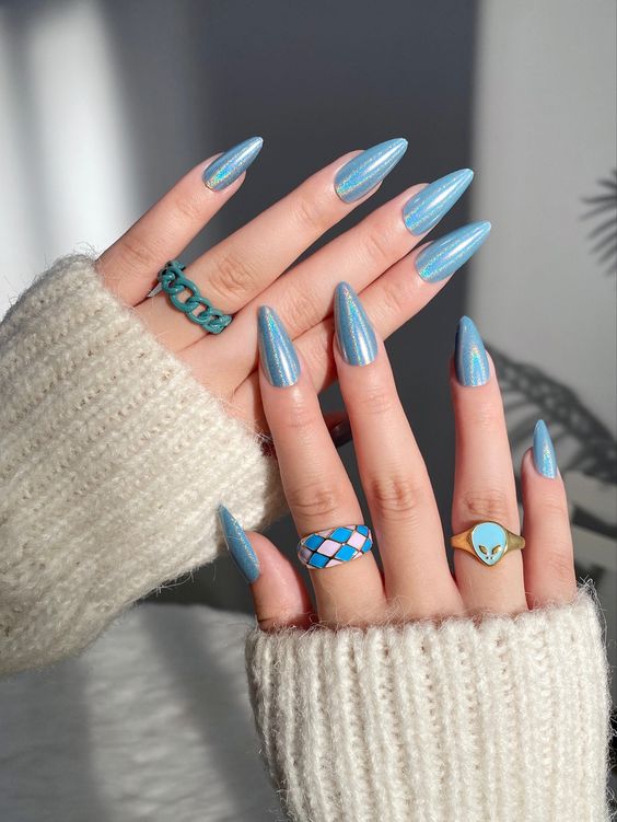 Mãos brancas sobrepostas com anéis coloridos e unha stiletto amendoada azul com glitter 