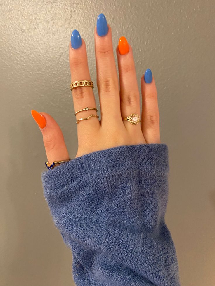 Mão branca com anéis dourados e unha laranja e azul 