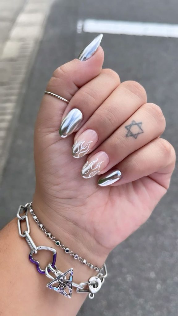 Mão branca curvada com dedo tatuado e unha prata metálica com decoração de chamas