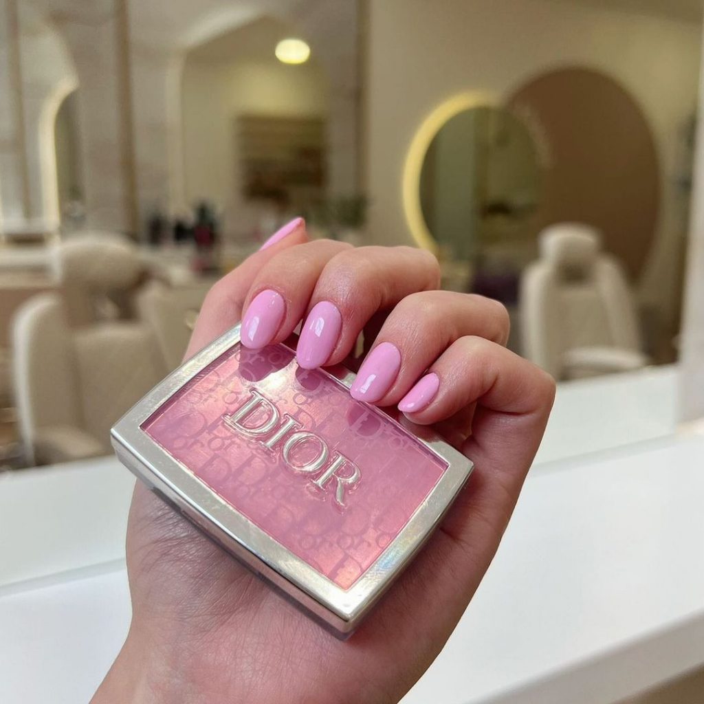 Mão branca curvada segundo um blush rosa da Dior, com unha rosa claro 