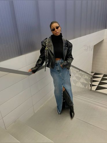 Moça jovem, magra e de pele escura. Ela usa jaqueta de couro preta, oculos de sol, blusa preta e saia longa jeans