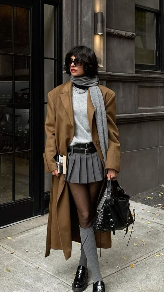 Mulher jovem, magra e de pele clara. Ela usa trench coat marrom, suéter cinza, saia cinza com dois cintos pretos, meia-calça preta, meia tres quartos cinza e mocassim preto.