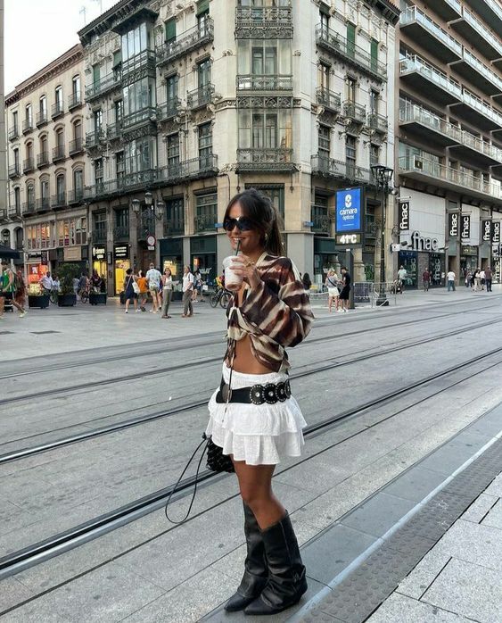 Mulher jovem e magra de cabelos castanhos. Ela usa blusa estampada, saia branca, cinto preto e bota de cano alto preta.