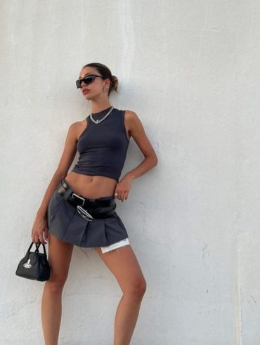 Mulher jovem, magra e de pele clara. Ela usa trench coat marrom, suéter cinza, saia cinza com dois cintos pretos, meia-calça preta, meia tres quartos cinza e mocassim preto.