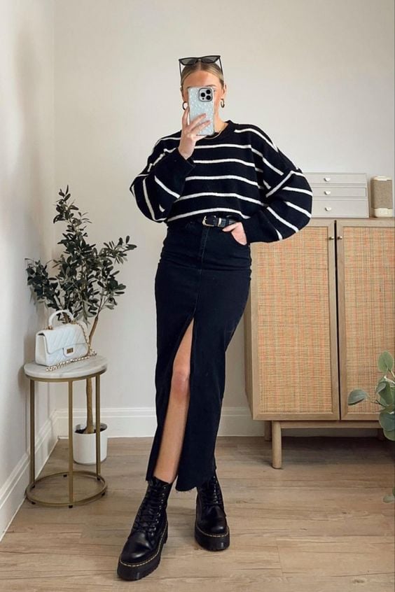 Mulher jovem, magra e de pele clara. Ela usa suéter listrado, saia longa jeans com cinto e coturno.