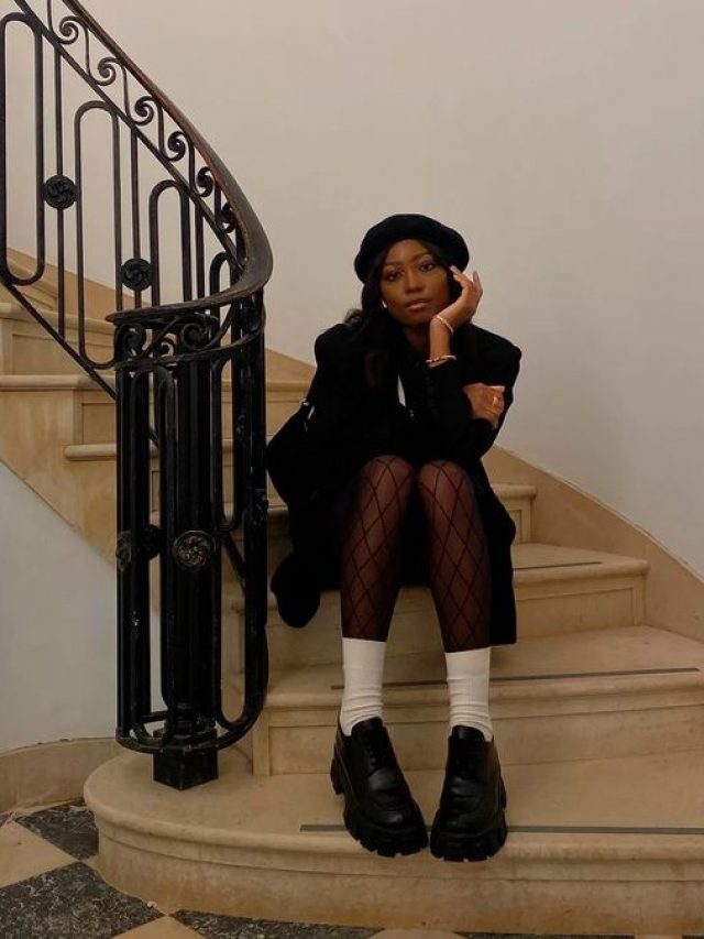 Moça jovem de pele escura. Ela está usando boina, sobretudo, meia-calça, mocassim e meia branca