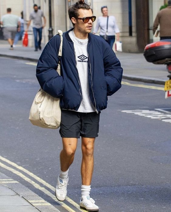 Harry Styles. Ele usa jaqueta azul, bermuda cinza e tênis Adidas Samba