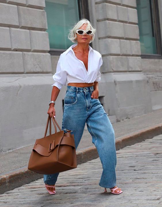 Mulher madura e de pele clara. Ela está usando camisa branca e calça jeans.