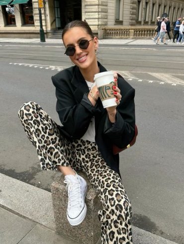 Mulher jovem de pele clara e cabelo escuro. Ela usa blazer preto, calça de oncinha e all star branco.