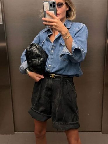Mulher jovem e magra. Ela tem pele clara e cabelo loiro curto. Ela veste camisa jeans de manga comprida e short jeans