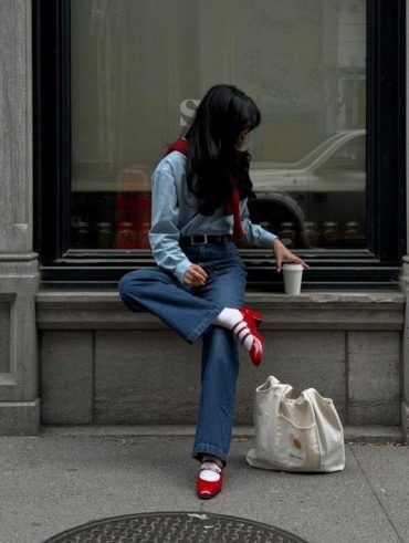 Moça jovem de cabelos escuros. Ela está usando camisa azul, cinto preto, calça jeans e sapato vermelho
