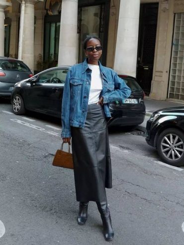 Mulher jovem, magra e de pele escura. Ela está vestindo jaqueta jeans azul, camiseta branca, saia de couro longa preta e botas de cano longo pretas.