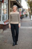 Mulher de meia idade, magra, de pele clara e cabelos curtos e grizalhos. Ela usa suéter listrado e calça jeans preta.