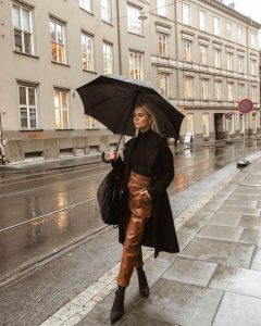 Mulher jovem e loira, segurando guarda-chuva , usando tricô preto, sobretudo preto e calça de couro marrom.