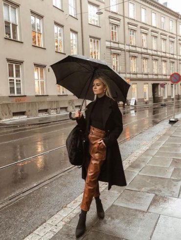 Mulher jovem e loira, segurando guarda-chuva , usando tricô preto, sobretudo preto e calça de couro marrom.