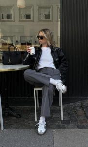 Mulher jovem, magra e loira, sentada com as pernas cruzadas e segurando um café, utilizando jaqueta de couro preta, blusa branca, calça de alfaiataria cinza e Adidas Samba.