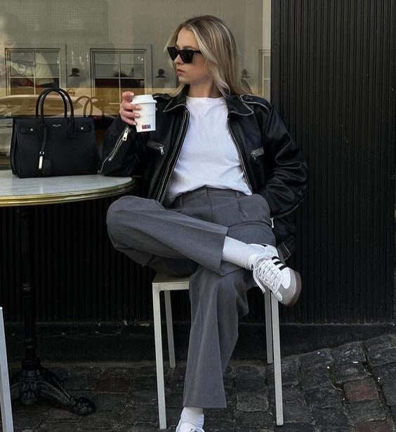 Mulher jovem, magra e loira, sentada com as pernas cruzadas e segurando um café, utilizando jaqueta de couro preta, blusa branca, calça de alfaiataria cinza e Adidas Samba.
