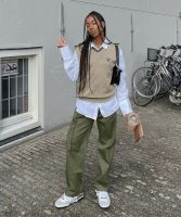 Mulher negra, usando colete em tricô na cor bege, com bolsa no ombro em cor preta, calça cargo em verde militar e tênis branco.