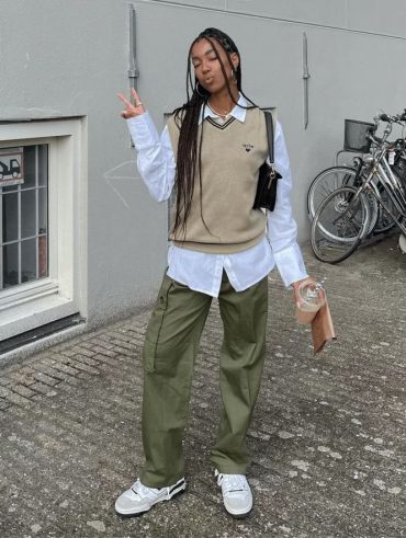 Mulher negra, usando colete em tricô na cor bege, com bolsa no ombro em cor preta, calça cargo em verde militar e tênis branco.