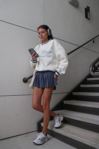 Mulher jovem, descendo escadas, usando fones de ouvidos e mexendo em celular, usando blusão em cor branca, saia de pregas em azul e tênis esportivo.