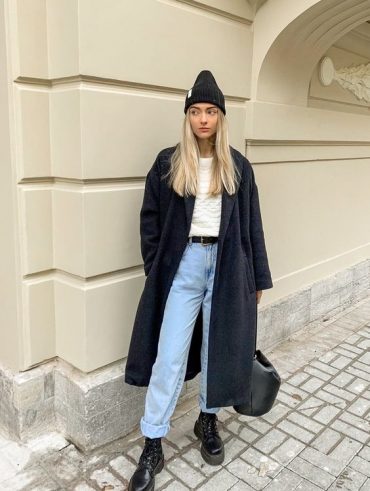 Mulher jovem e loira, usando touca preta, blusa branca, calça jeans com cinto preto, e sobretudo preto,