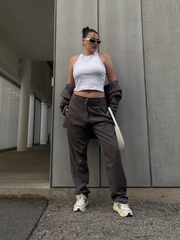 Mulher jovem usando coque no cabelo, óculos de sol, usando regata branca, calça cinza e tênis.