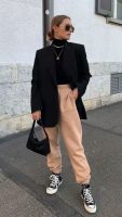 Mulher com cabelo preso em coque, usando óculos de sol, blazer preto, calça de moletom bege e usando all star de botinha preto.