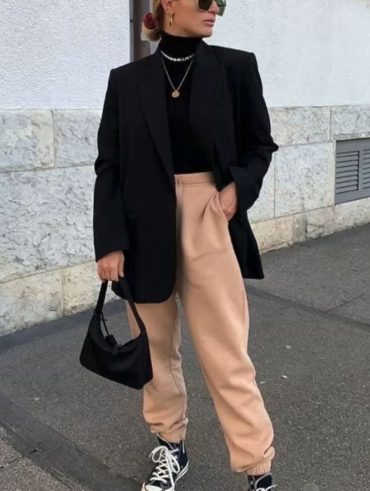Mulher com cabelo preso em coque, usando óculos de sol, blazer preto, calça de moletom bege e usando all star de botinha preto.