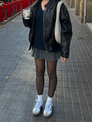 Mulher vestindo jaqueta preta, segurando um copo de café, usando saia cinza e meia-calça preta com Adidas Samba.
