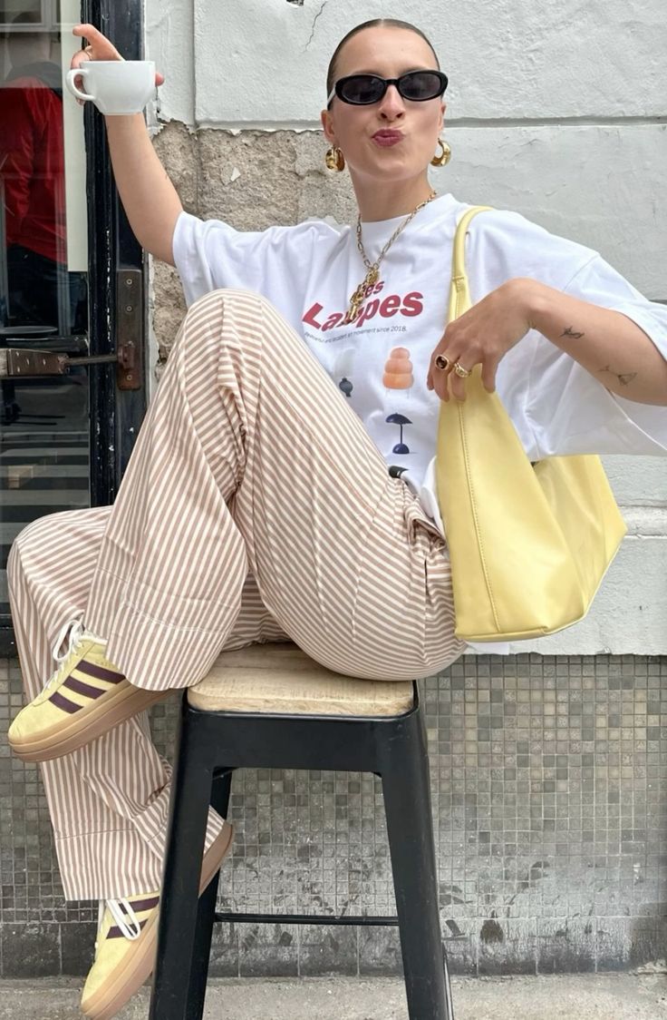 Pessoa sentada usando camiseta oversized branca com estampa gráfica, calça listrada bege e branca, tênis amarelos com listras roxas, bolsa amarela e óculos de sol pretos. Look descontraído e moderno, acessórios incluem brincos de argola dourados e anéis.