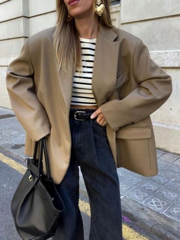 Mulher branca usando blazer oversized, brinco maximalista em formato de coração, blusa listrada, calça jeans e maxi bag preta